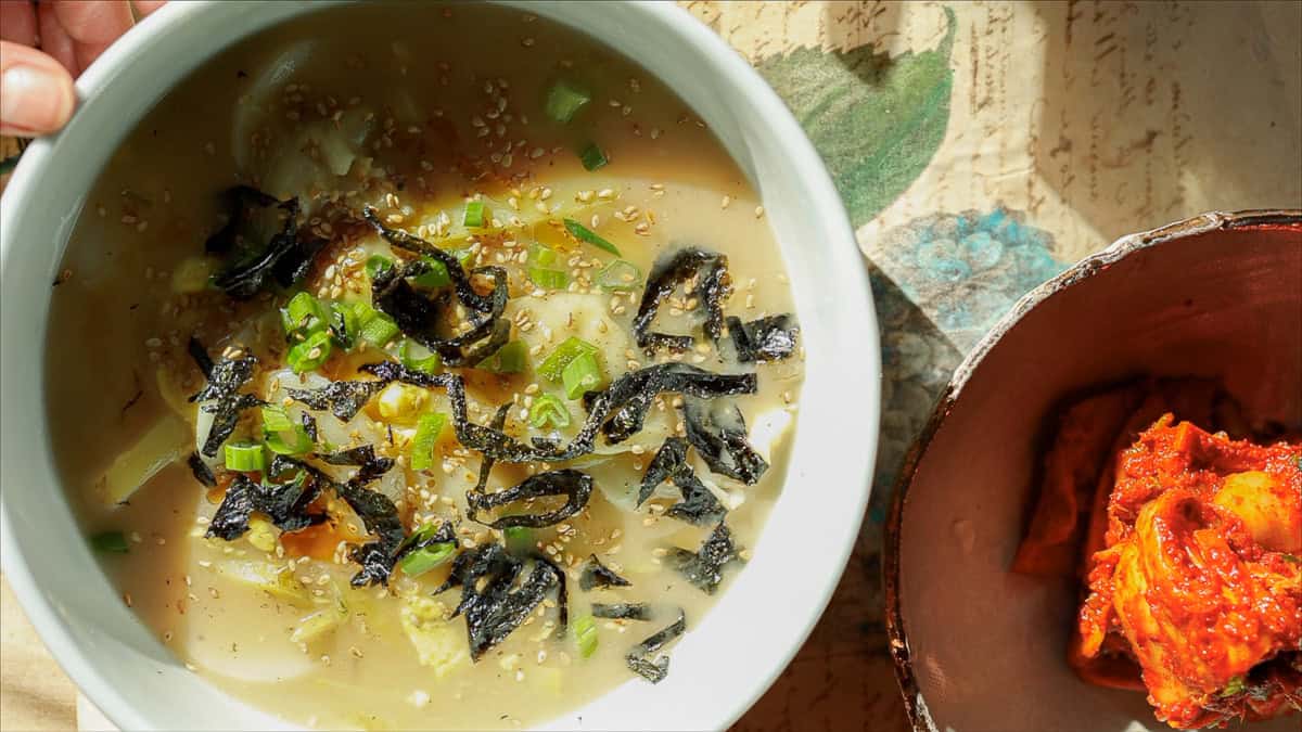 A bowl of steaming vegan rice cake soup with sliced vegetables, sprinkled with seaweed, sesame seeds, and green onions, sits on a textured surface. Beside it is a dish of vibrant red kimchi, partially visible.